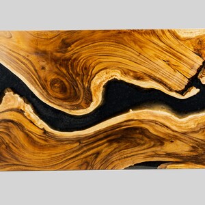 May include: A close-up of a wooden table top with a black resin river running through it. The wood has a rich brown color with natural grain patterns.