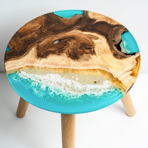 May include: Round wooden stool with three legs. The top of the stool is made of wood and has a blue, white, and brown epoxy resin design that resembles an ocean wave.