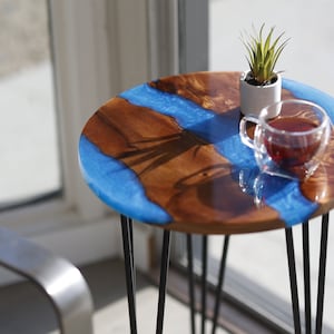 May include: A round wooden table with a blue epoxy resin top. The table has black metal hairpin legs and a small potted plant and a glass of tea on top.