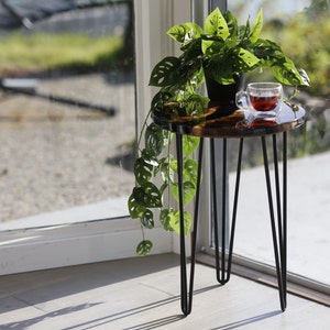 May include: A small, round, brown and black epoxy resin table with a black metal hairpin leg base. A potted plant with green leaves and a glass of red liquid sits on the table.
