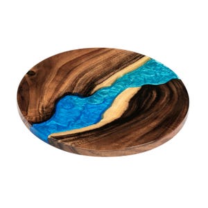 May include: Oval wooden serving board with a river of blue resin. The wood grain is a rich brown, and the resin has a swirling, water-like appearance. The board is a decorative and functional item.