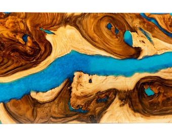 May include: A close-up of a wooden table top with a blue resin inlay. The wood has a natural brown and light brown color with a swirling grain pattern. The resin is a bright blue color and flows through the wood in a river-like shape.