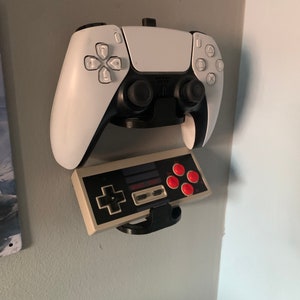 May include: A white PlayStation 5 controller and a grey Nintendo Entertainment System controller mounted on a wall with a black holder.