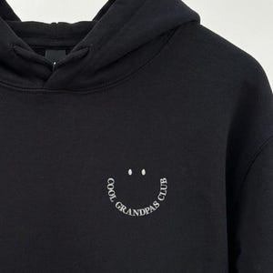 Puede incluir: Sudadera con capucha negra con cordón y un gráfico de carita sonriente blanca que dice "COOL GRANDPAS CLUB". La sudadera está colgada en una percha de madera.
