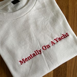Weißes T-Shirt mit dem in Rot gestickten Schriftzug "Mentally On A Yacht". Das Shirt hat einen Rundhalsausschnitt und kurze Ärmel. Der Stoff ist 100% Baumwolle.