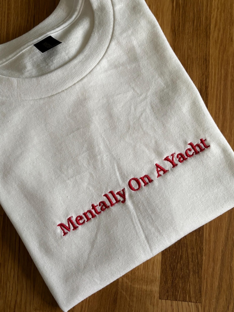 Weißes T-Shirt mit dem in Rot gestickten Schriftzug "Mentally On A Yacht". Das Shirt hat einen Rundhalsausschnitt und kurze Ärmel. Der Stoff ist 100% Baumwolle.
