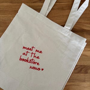 Könnte beinhalten: Beige Canvas-Tragetasche mit langen Henkeln. Der Beutel hat einen roten, gestickten Text: "meet me at the bookstore xoxo ♥". Die Tragetasche ist aus robustem Stoff gefertigt und eignet sich zum Tragen von Büchern und anderen Gegenständen.