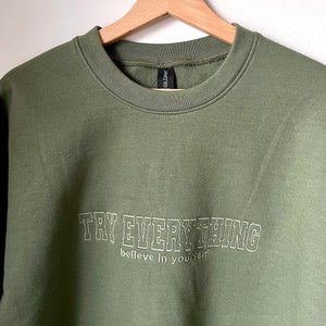 Könnte beinhalten: Ein grüner Pullover mit dem Text "TRY EVERYTHING believe in yourself" in weißen umrandeten Buchstaben.
