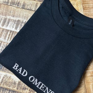 Ein gefaltetes schwarzes T-Shirt mit dem gestickten Text "BAD OMENS" in Weiß. Das Shirt besteht aus einem weichen Material und hat einen Rundhalsausschnitt. Das Gildan-Markenetikett ist sichtbar.