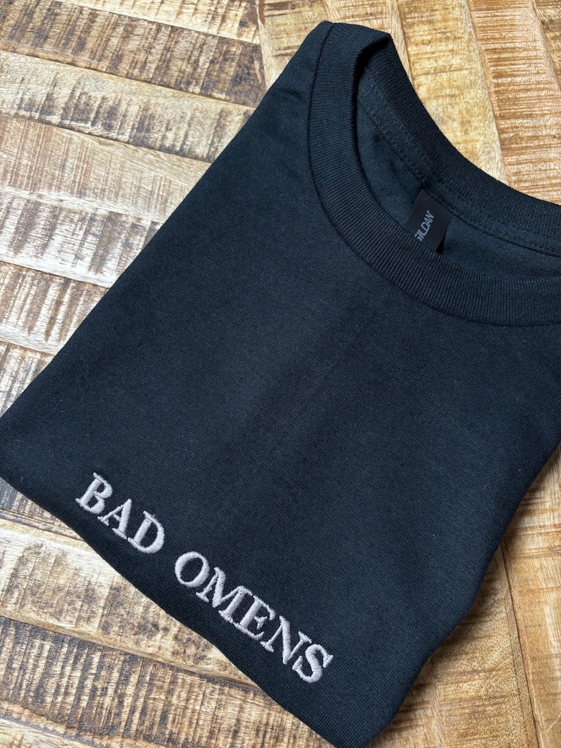 Ein gefaltetes schwarzes T-Shirt mit dem gestickten Text "BAD OMENS" in Weiß. Das Shirt besteht aus einem weichen Material und hat einen Rundhalsausschnitt. Das Gildan-Markenetikett ist sichtbar.