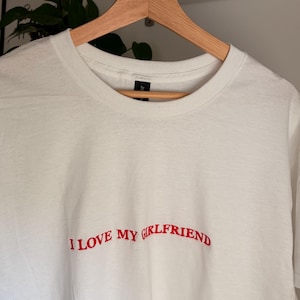 Könnte beinhalten: Ein weißes Kurzarm-T-Shirt mit dem in Rot gestickten Text "I LOVE MY GIRLFRIEND" mittig auf der Brust. Das Kleidungsstück hat einen klassischen Rundhalsausschnitt und hängt an einem Holzkleiderbügel, was ein schlichtes Design zeigt.