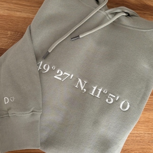 Op de afbeelding: Een saliegroen hoodie met witte geborduurde tekst die "49°27'N, 11°5'0" luidt.