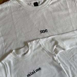 wei weiße T-Shirts, eines für Erwachsene und eines für Babys, mit den Wörtern "me" und "mini me" in Schwarz gestickt. Das T-Shirt für Erwachsene hat einen Rundhalsausschnitt, und das Baby-T-Shirt hat Druckknöpfe an der Schulter. Die Shirts sind aus weichem, schlichtem 100% Baumwollstoff.