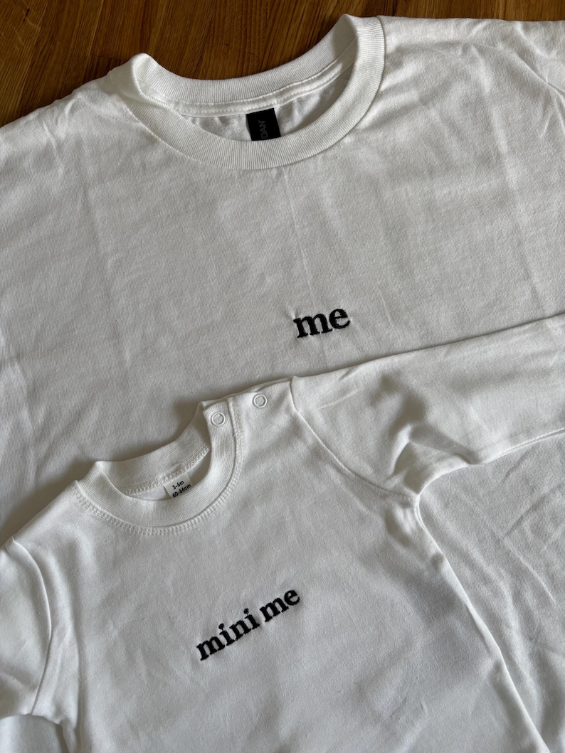 wei weiße T-Shirts, eines für Erwachsene und eines für Babys, mit den Wörtern "me" und "mini me" in Schwarz gestickt. Das T-Shirt für Erwachsene hat einen Rundhalsausschnitt, und das Baby-T-Shirt hat Druckknöpfe an der Schulter. Die Shirts sind aus weichem, schlichtem 100% Baumwollstoff.