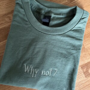 Ein salbeigrünes T-Shirt mit dem gestickten Schriftzug "Why not?" in einer Schreibschrift. Das Shirt ist sauber gefaltet und zeigt den Rundhalsausschnitt und die weiche Textur des Stoffes. Das Shirt liegt auf einer Holzoberfläche.