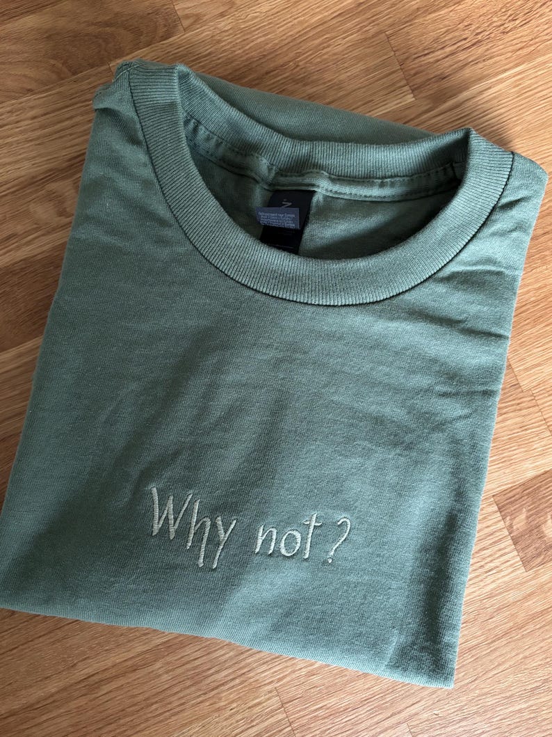 Ein salbeigrünes T-Shirt mit dem gestickten Schriftzug "Why not?" in einer Schreibschrift. Das Shirt ist sauber gefaltet und zeigt den Rundhalsausschnitt und die weiche Textur des Stoffes. Das Shirt liegt auf einer Holzoberfläche.