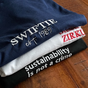 May include: A stack of three t-shirts with different embroidered designs. The top shirt is navy blue with white embroidery that says "SWIFTIE est. 1989". The middle shirt is white with red embroidery that says "ZIRKU". The bottom shirt is black with white embroidery that says "sustainability is not a crime".