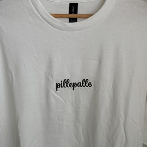Weißes T-Shirt mit dem in Coffee Brown gestickten Schriftzug "pillepalle". Das Shirt hat einen Rundhalsausschnitt und kurze Ärmel. Der Stoff ist 100% Baumwolle.