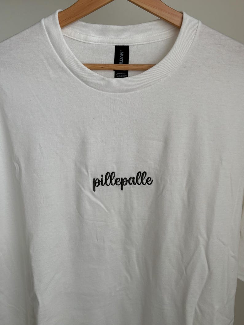Weißes T-Shirt mit dem in Coffee Brown gestickten Schriftzug "pillepalle". Das Shirt hat einen Rundhalsausschnitt und kurze Ärmel. Der Stoff ist 100% Baumwolle.