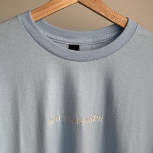 Hellblaues T-Shirt mit dem gestickten Schriftzug "you're so golden" in Schreibschrift. Das T-Shirt hängt an einem Holzkleiderbügel. Es hat einen Rundhalsausschnitt und kurze Ärmel.