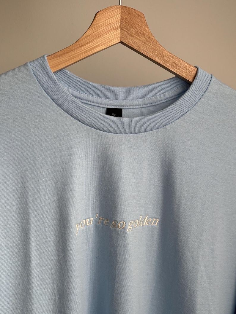 Hellblaues T-Shirt mit dem gestickten Schriftzug "you're so golden" in Schreibschrift. Das T-Shirt hängt an einem Holzkleiderbügel. Es hat einen Rundhalsausschnitt und kurze Ärmel.