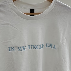 Weißes T-Shirt mit Rundhalsausschnitt und dem gestickten Text "IN MY UNCLE ERA" in Hellblau. Das Shirt hängt an einem Holzkleiderbügel vor weißem Hintergrund.