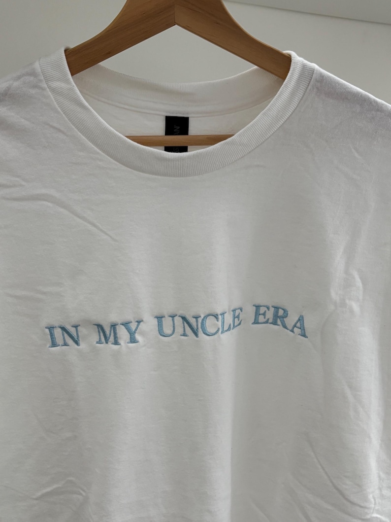 Weißes T-Shirt mit Rundhalsausschnitt und dem gestickten Text "IN MY UNCLE ERA" in Hellblau. Das Shirt hängt an einem Holzkleiderbügel vor weißem Hintergrund.