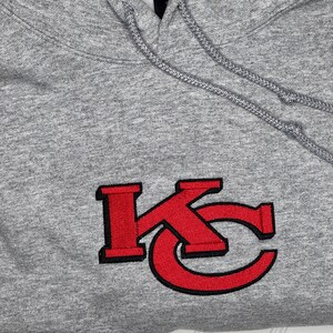 K&ouml;nnte beinhalten: Grauer Kapuzenpullover mit einem roten gestickten "KC"-Logo.