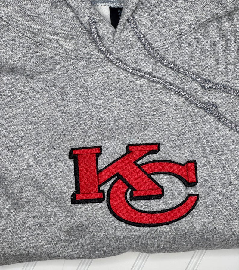 K&ouml;nnte beinhalten: Grauer Kapuzenpullover mit einem roten gestickten "KC"-Logo.