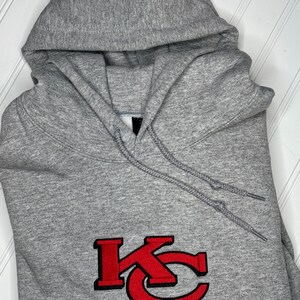 K&ouml;nnte beinhalten: Grauer Kapuzenpullover mit einem roten gestickten "KC"-Logo.