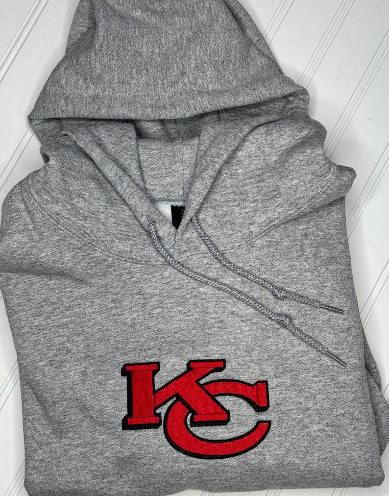K&ouml;nnte beinhalten: Grauer Kapuzenpullover mit einem roten gestickten "KC"-Logo.