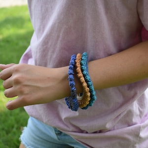 Könnte beinhalten: Eine Person trägt ein rosa Shirt mit drei gehäkelten Armbändern am Handgelenk. Die Armbänder sind blau, türkis und orange.