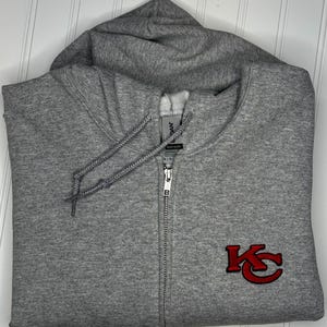 K&ouml;nnte beinhalten: Ein grauer Kapuzenpullover mit Rei&szlig;verschluss und einem roten gestickten "KC"-Logo auf der Vorderseite.