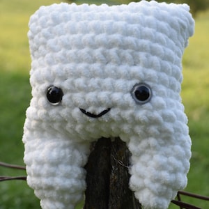 Peut inclure: Un jouet en peluche blanc en crochet en forme de dent avec un visage souriant et des yeux noirs. Le jouet est assis sur un poteau de clôture en bois.