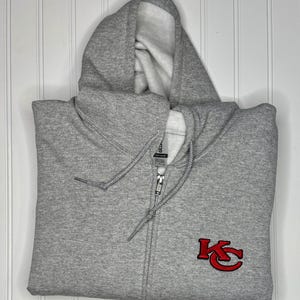 K&ouml;nnte beinhalten: Ein grauer Kapuzenpullover mit Rei&szlig;verschluss und einem roten gestickten "KC"-Logo auf der Vorderseite.