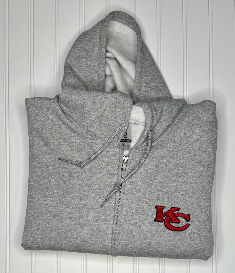 K&ouml;nnte beinhalten: Ein grauer Kapuzenpullover mit Rei&szlig;verschluss und einem roten gestickten "KC"-Logo auf der Vorderseite.