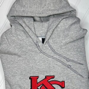K&ouml;nnte beinhalten: Ein grauer Kapuzenpullover mit einem roten gestickten "KC"-Logo.