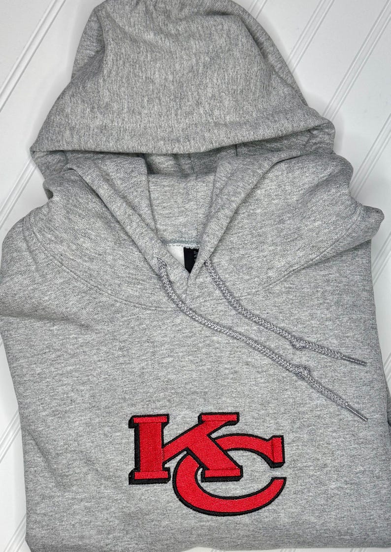 K&ouml;nnte beinhalten: Ein grauer Kapuzenpullover mit einem roten gestickten "KC"-Logo.