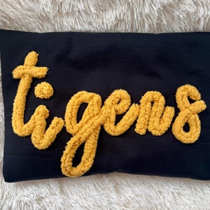 May include: Black pillow with the word "Tigers" in yellow, textured, raised lettering. The pillow is rectangular and sits on a white, fluffy surface. The yellow lettering is in a script font.