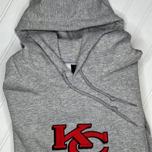 K&ouml;nnte beinhalten: Ein grauer Kapuzenpullover mit einem roten gestickten "KC"-Logo auf der Vorderseite.