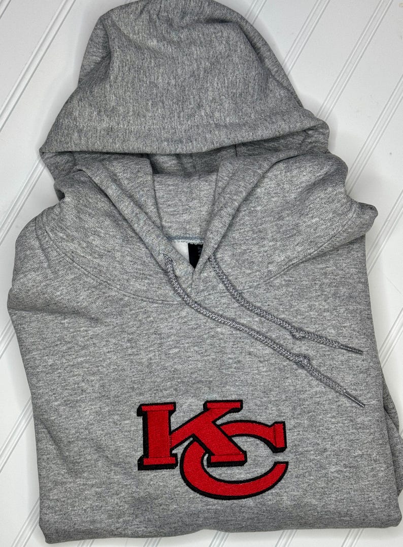K&ouml;nnte beinhalten: Ein grauer Kapuzenpullover mit einem roten gestickten "KC"-Logo auf der Vorderseite.