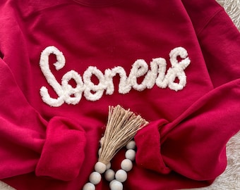 May include: A red sweatshirt with the word "Sooners" in white, fluffy letters across the chest. The sleeves are folded, and a beaded tassel is draped over the front. The sweatshirt is on a white, fluffy surface.