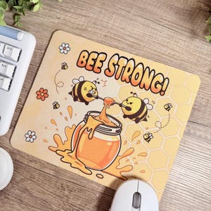 Könnte beinhalten: Rechteckiges Mauspad mit einem Cartoon-Bienen-Design. Das Mauspad zeigt den Text "BEE STRONG!" über einem Honigglas, das von zwei wütenden Bienen angehoben wird. Der Hintergrund ist hellgelb mit einem Wabenmuster.
