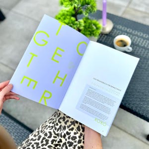 Könnte beinhalten: Ein offenes Buch mit dem Wort "TOGETHER" in leuchtend grünen Buchstaben auf einer hellvioletten Seite. Die gegenüberliegende Seite hat Text auf Deutsch. Eine Tasse Kaffee und eine kleine grüne Pflanze sind im Hintergrund.