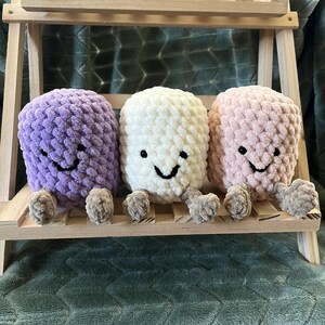 Peut inclure: Trois jouets en peluche souriants, violets, blancs et roses, chacun avec de petits pieds attachés. Ils sont disposés sur une petite étagère en bois, avec une couverture verte en arrière-plan. Les jouets ont des sourires brodés noirs.