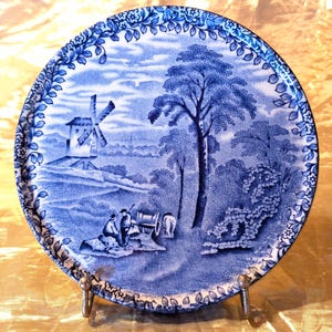 May include: A blue and white ceramic plate with a windmill and a tree in a landscape scene. The plate has a decorative border of blue leaves and flowers.