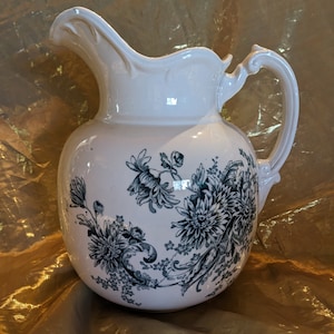 May include: White ceramic pitcher with a black floral design. The pitcher has a wide mouth and a handle.