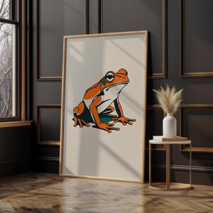 May include: A framed art print featuring a stylized illustration of a frog. The frog is depicted in shades of orange, green, and white against a cream-colored background. The artwork is displayed in a room with dark walls and a wooden floor.