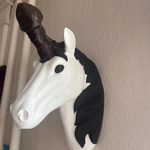 Op de afbeelding: Een witte en zwarte eenhoofdige muursculptuur met een bruine hoorn. De sculptuur is bevestigd aan een lichtbeige muur. De eenhoorn heeft zwart haar en een wit gezicht.