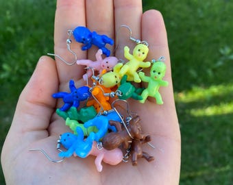 Op de afbeelding: Een verzameling kleurrijke baby-poppen oorbellen in verschillende kleuren, waaronder blauw, roze, geel, groen, oranje en bruin. Elke oorbel heeft een kleine plastic baby-poppenbedel die aan een zilverkleurige haak hangt.